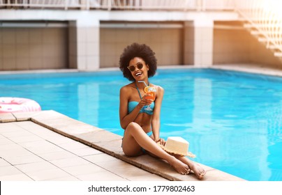 Young black woman in swimsuit relaxing with summer cocktail near pool at tropical resort