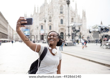 Young black woman taking selfie with smart phone hand hold with Milan cathedral in background, smiling - happiness, traveler, technology concept