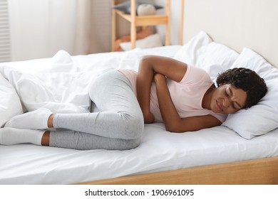 Young black woman touching her belly, laying in bed, side view, copy space. Sad african american lady suffering from abdominal pain, hugging her tummy and crying, home interior