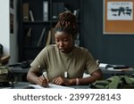 Young black woman in t-shirt making notes in notepad after teacher at military training lesson while sitting by desk in classroom