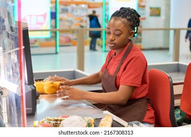 Young Black Woman In Uniform Scanning Food Products And Putting Them On Other Side Of Counter While Sitting By Cashier In Supermarket