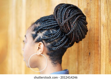 Young Black Woman With An Updo Braided Bun Hairstyle And Silver Hoop Earrings Stands Outside Against A Wooden Wall                               