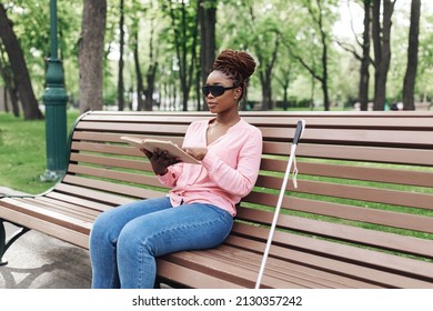 Young Black Woman With Vision Disability Sitting On Bench In City Park, Reading Braille Book Outdoors, Copy Space. Millennial Blind Lady In Dark Glasses Studying, Using Textbook Outside