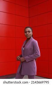 Young Black Woman Wearing Corporate Formal Purple Suit For Looking Smart. Urban Red Wall Background, Posing Outside. Pink Make Up, Braids Hairstyle. Going To Her Workplace, Formal Gatherings, Date