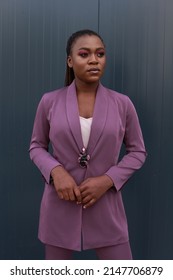 Young Black Woman Wearing Corporate Formal Purple Suit For Looking Smart. Urban Grey Wall Background, Posing Outside. Pink Make Up, Braids Hairstyle. Going To Her Workplace, Formal Gatherings, Date