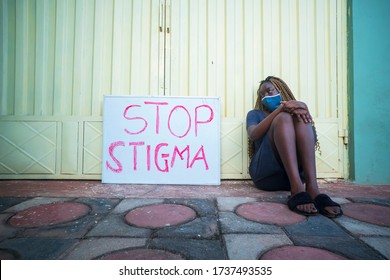 Young Black Woman Wearing A Locally Made Mask, Sitting Outdoors With Campaign Sign On Stop Covid-19 (corona Virus) Stigma - Concept On Infected People With Covid-19 And Stigmatization( Social Stigma )