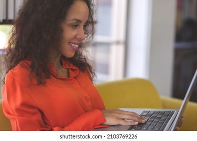 Young Black Woman Wearing Orange Shirt Smiling And Using Laptop For Shopping Online, Leaning Online, And Video Call On Sofa At Home. Business Woman Work From Home In Spreading Of Coronavirus Covid-19.