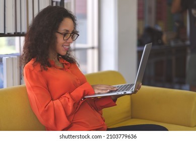 Young Black Woman Wearing Orange Shirt Smiling And Using Laptop For Shopping Online, Leaning Online, And Video Call On Sofa At Home. Business Woman Work From Home In Spreading Of Coronavirus Covid-19.