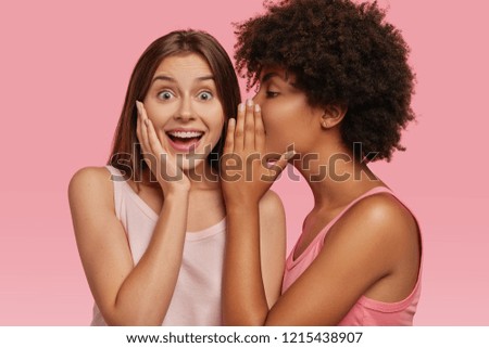 Young black woman whispers secret to her Caucasian friend, gossip together and spread rumours. Emotional European girl feels amazed to hear confidential information from companion. Secrecy, gossiping