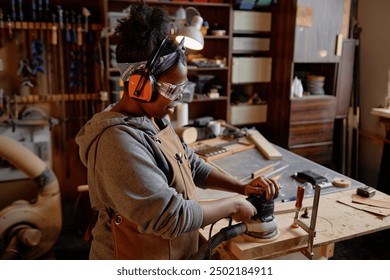 Young Black woman in workshop industriously sanding wood with protective gear surrounded by woodworking tools and materials - Powered by Shutterstock