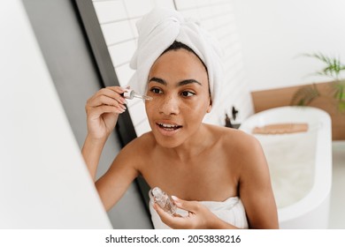 Young Black Woman Wrapped In Towels Applying Face Serum At Bathroom