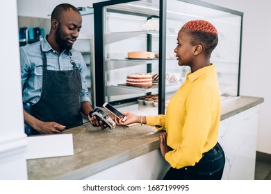 Young Black Woman In Yellow Shirt Making Payment With Mobile Phone On Payment Terminal In Cafe Standing Before Counter With Cakes And Desserts