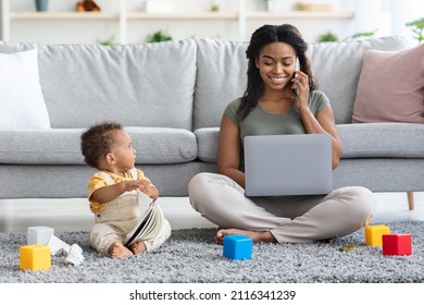 Young Black Working Mom Freelancing At Home And Taking Care About Baby, Smiling African American Woman Using Laptop And Talking On Cellphone While Her Infant Child Playing Around, Copy Space