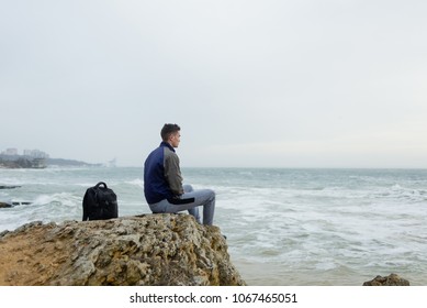 young black-haired guy with a backpack sitting on a rock above the sea and looking out into the distance