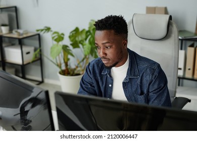 Young blackman in casual clothes sitting in front of computer monitors and looking at data on screens during work in openspace bureau