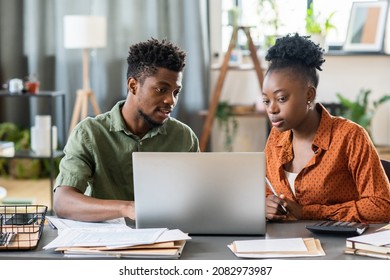 Young blackman sitting at table with bills and document and examining online tax form with girlfriend on laptop