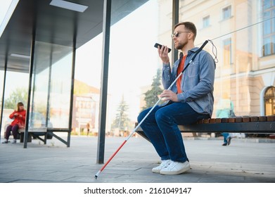 Young blind man with smartphone sitting on bench in park in city, calling - Shutterstock ID 2161049147