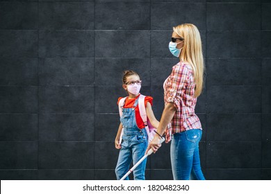 Young blind mother walking with her little daughter on city street. They wearing face protective masks. Back to school and new coronavirus lifestyle concept.