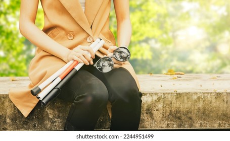 Young blind woman with vision disabilities takes off sunglasses protects eyesight and holds blind cane sitting calmly on concrete platform relaxing in park : Self care blind disabled woman concept. - Shutterstock ID 2244951499