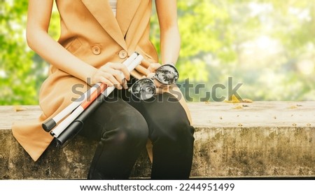 Young blind woman with vision disabilities takes off sunglasses protects eyesight and holds blind cane sitting calmly on concrete platform relaxing in park : Self care blind disabled woman concept.