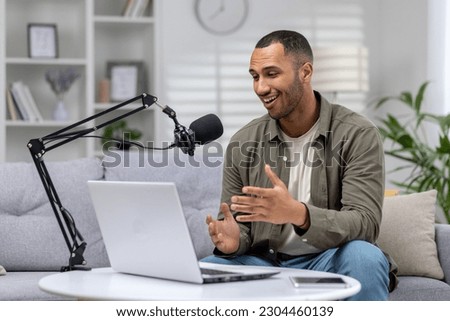 Young blogger at home on couch with laptop recording online podcast, man with professional microphone streaming from living room, hispanic cheerfully telling success stories to subscribers. [[stock_photo]] © 