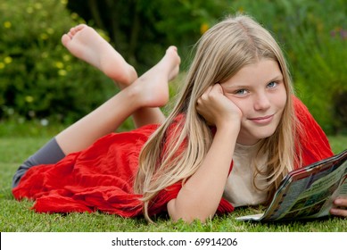 Young blond blue-eyed teenage girl lying on the grass on the garden and reading a teen magazine.