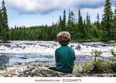 A young blond boy sitting on rocky riverbank, gazing at the rushing rapids of Storån River, surrounded by a dense forest on a warm summer day in the Nordic Wilderness - Powered by Shutterstock