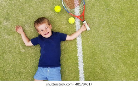 A young blond boy smiling and laying on a tennis court, with racket and balls.