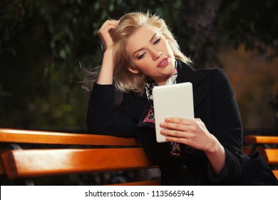 Young blond business woman using digital tablet computer sitting on the bench Stylish fashion female model wearing classic black coat  - Powered by Shutterstock