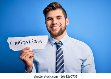 Young blond businessman with beard and blue eyes holding paper with capitalism message with a happy face standing and smiling with a confident smile showing teeth