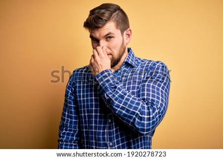 Young blond businessman with beard and blue eyes wearing shirt over yellow background smelling something stinky and disgusting, intolerable smell, holding breath with fingers on nose. Bad smell