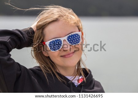 Young blond caucasian girl wearing American flag themed sunglasses holding on to her hair and smiling on a windy cloudy day.