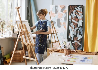 Young blond craftswoman in apron painting new masterpiece on canvas in studio of contemporary art while standing in front of easel - Powered by Shutterstock