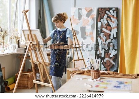 Young blond craftswoman in apron painting new masterpiece on canvas in studio of contemporary art while standing in front of easel