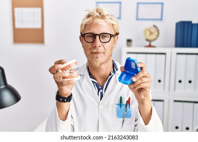 Young blond dentist man working at dentist clinic holding brackets and aligner relaxed with serious expression on face. simple and natural looking at the camera. 