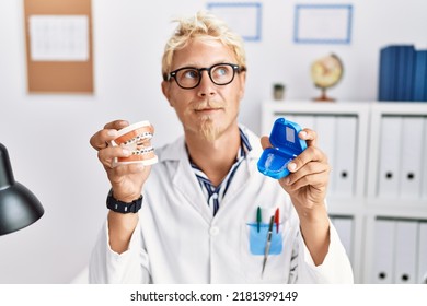 Young blond dentist man working at dentist clinic holding brackets and aligner smiling looking to the side and staring away thinking. 