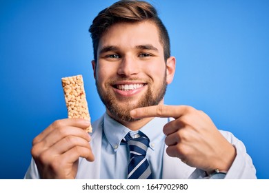 Young Blond Doctor Man With Beard And Blue Eyes Wearing Coat Eating Granola Bar Very Happy Pointing With Hand And Finger