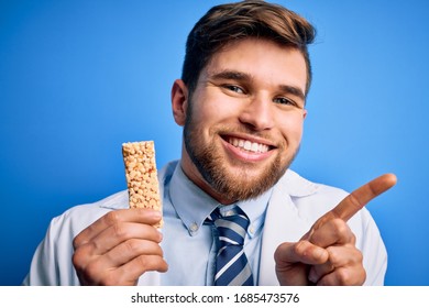 Young Blond Doctor Man With Beard And Blue Eyes Wearing Coat Eating Granola Bar Very Happy Pointing With Hand And Finger To The Side