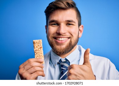 Young Blond Doctor Man With Beard And Blue Eyes Wearing Coat Eating Granola Bar Happy With Big Smile Doing Ok Sign, Thumb Up With Fingers, Excellent Sign