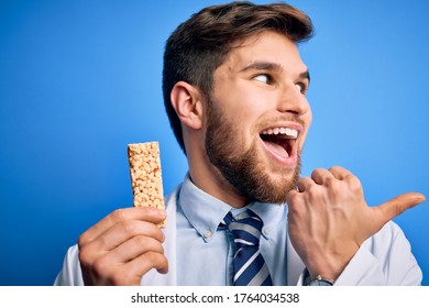 Young Blond Doctor Man With Beard And Blue Eyes Wearing Coat Eating Granola Bar Pointing And Showing With Thumb Up To The Side With Happy Face Smiling