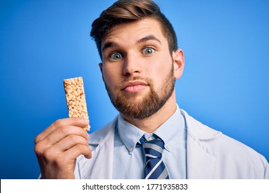 Young Blond Doctor Man With Beard And Blue Eyes Wearing Coat Eating Granola Bar With A Confident Expression On Smart Face Thinking Serious