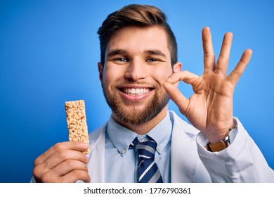 Young Blond Doctor Man With Beard And Blue Eyes Wearing Coat Eating Granola Bar Doing Ok Sign With Fingers, Excellent Symbol