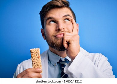 Young Blond Doctor Man With Beard And Blue Eyes Wearing Coat Eating Granola Bar Serious Face Thinking About Question, Very Confused Idea