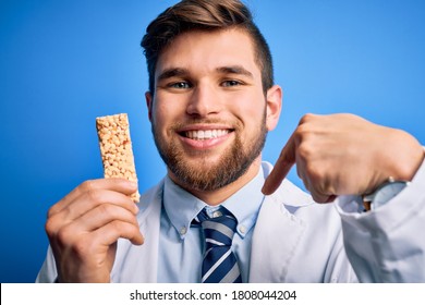 Young Blond Doctor Man With Beard And Blue Eyes Wearing Coat Eating Granola Bar With Surprise Face Pointing Finger To Himself