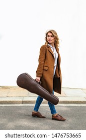 Young blond female musician in brown coat and jeans with violin in leather case in hand goes through city near white building at autumn or spring day. Music, hobby and art concept