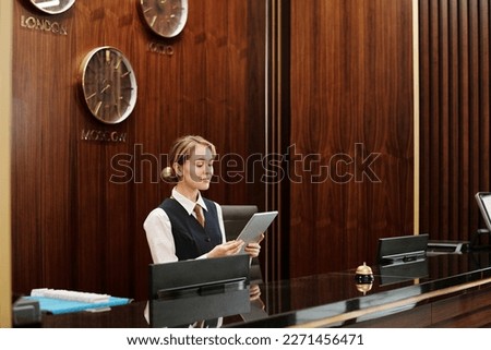 Young blond female receptionist in uniform looking at tablet screen while standing by counter in lounge of modern hotel and networking