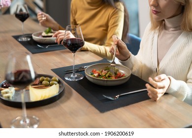 Young blond female in smart casualwear sitting by served table against her friend and having vegetable salad, red wine, cheese and olives