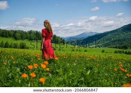 A young blond girl wearing red dress stay on a green lawn, and looking downhill over valley above cloud in mountains. Back view. Green valley high on the mountains