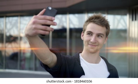 Young Blond Guy Making Selfie On Background Of Glass Building, Casual Guy With Phone Lens Flare. Portrait Of Smiling Guy Making Selfie On Background Of Business Building