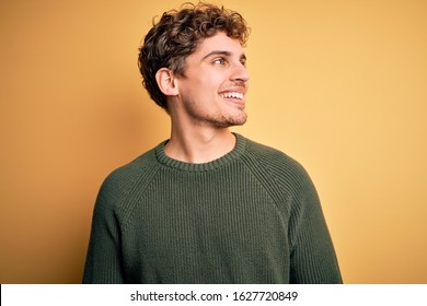 Young blond handsome man with curly hair wearing green sweater over yellow background looking away to side with smile on face, natural expression. Laughing confident. - Powered by Shutterstock